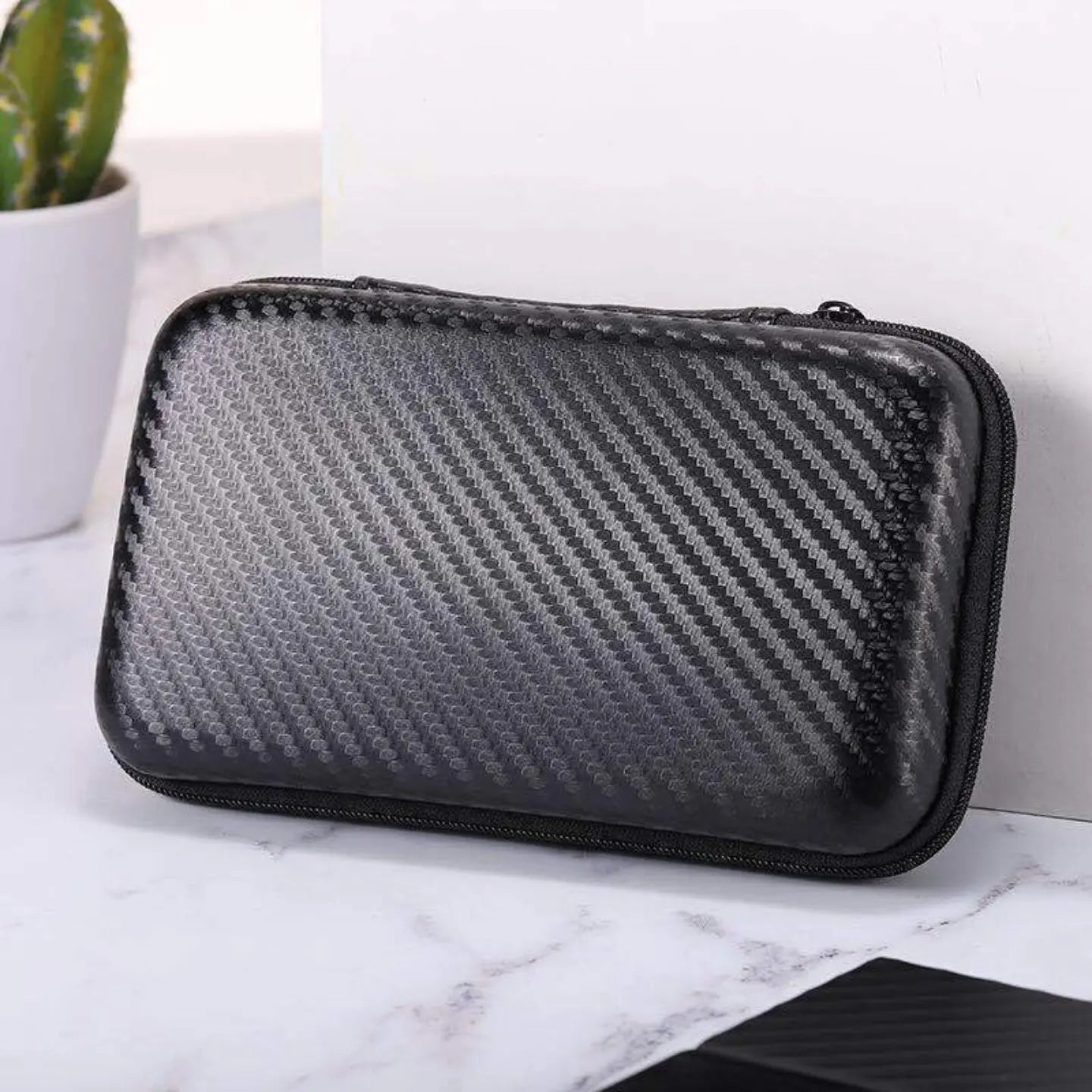 Black and gray carbon fiber patterned case on a marble surface with a plant in the background